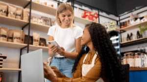 A imagem mostra duas mulheres interagindo sorridentes no interior de uma loja de produtos naturais ou empório comercial.