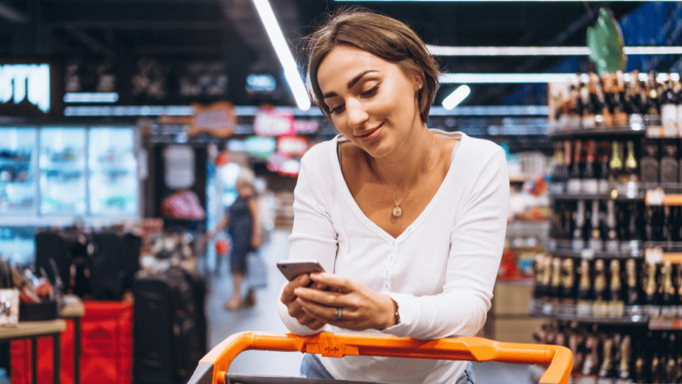 A imagem mostra uma mulher sorridente usando um smartphone enquanto faz compras em um supermercado.